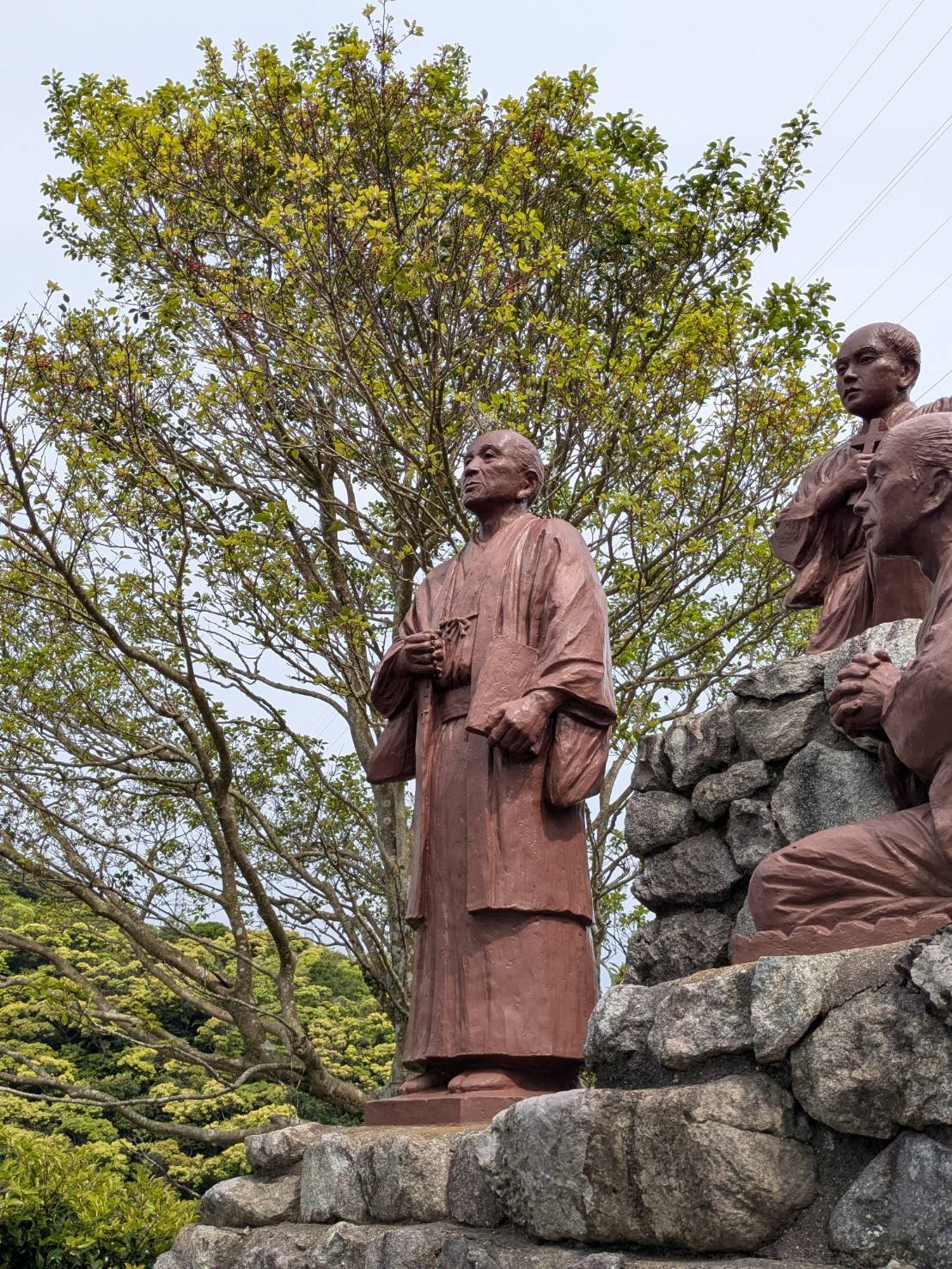 A standing statue with hands joined in prayer, surrounded by trees